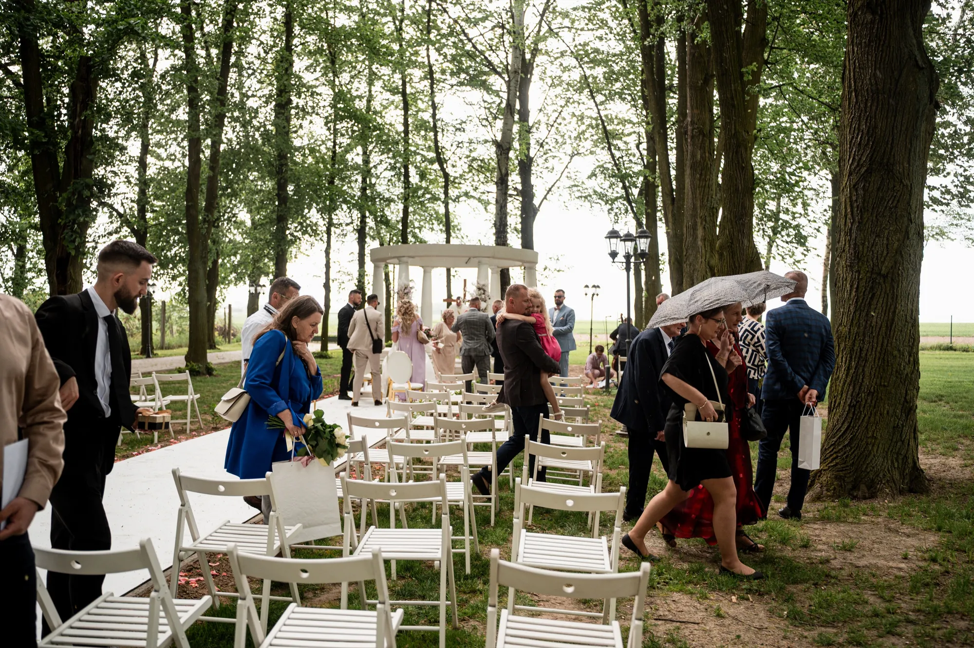 Goście w deszczu idą między drzewami, jedna z pań trzyma parasol, na trawie stoją białe krzesła, w tle pergola