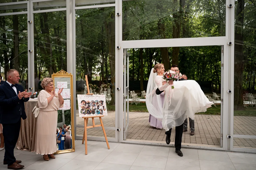 Rafał niesie Julię na rękach przez szklane drzwi sali, goście klaszczą, obok stoi tablica ze zdjęciami