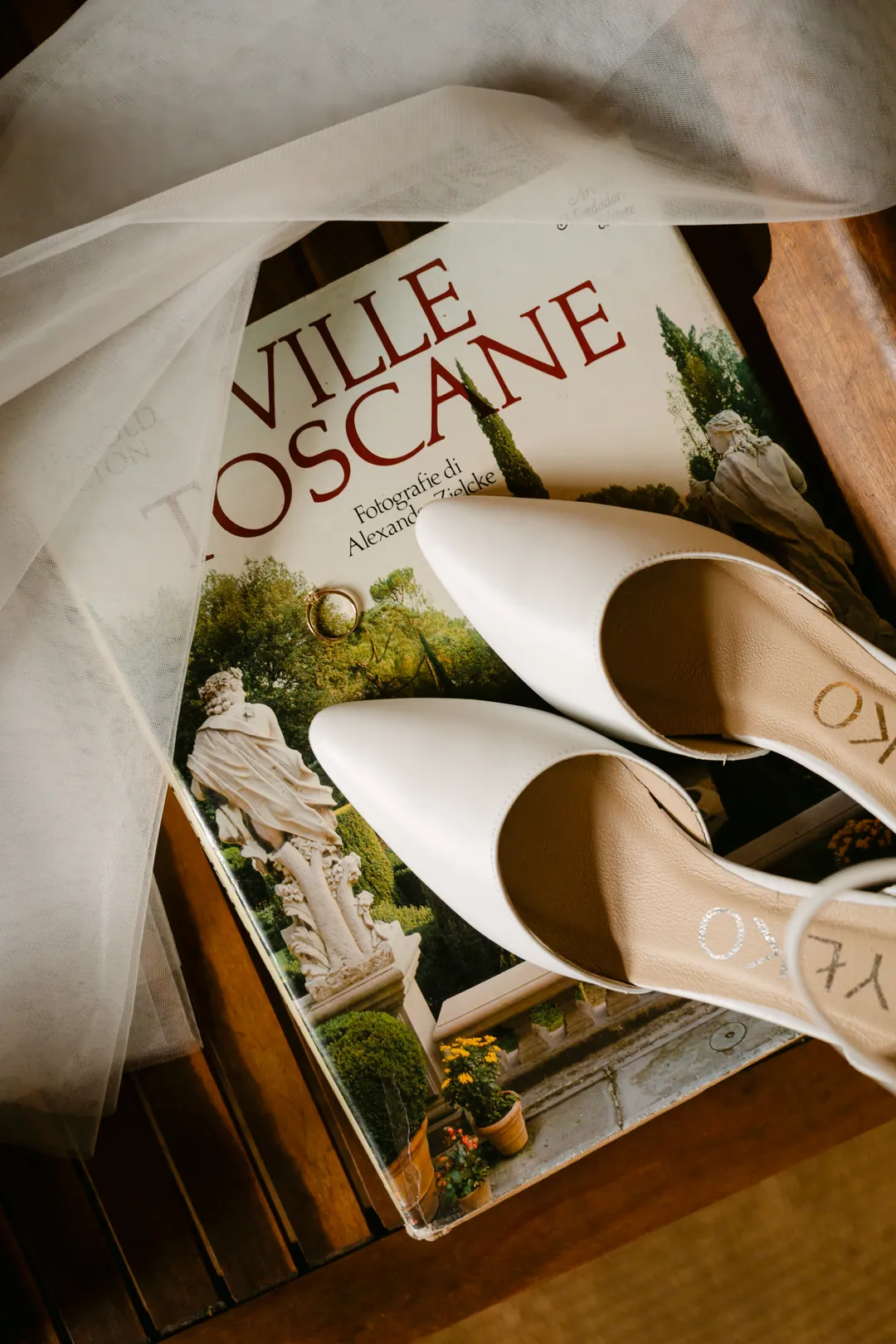 Białe buty i tiulowy welon na książce „Ville Toscane”, na okładce złota obrączka