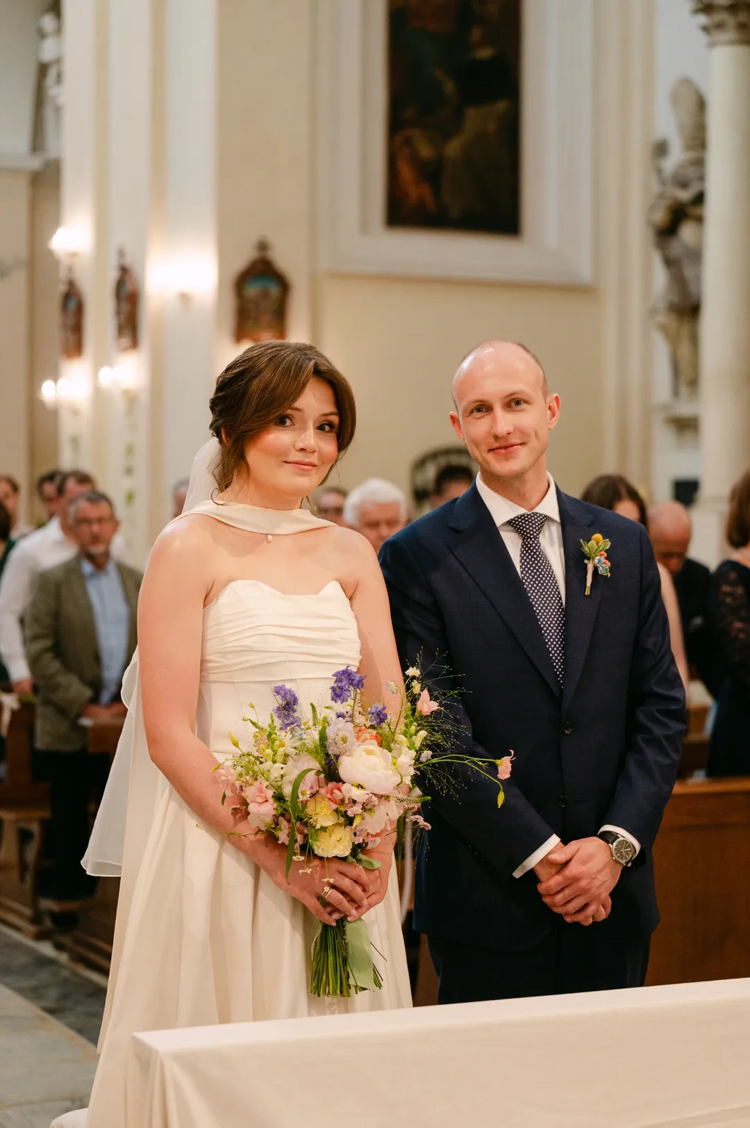 Kinga z bukietem polnych kwiatów i Mateusz w granatowym garniturze stoją w kościele, za nimi goście