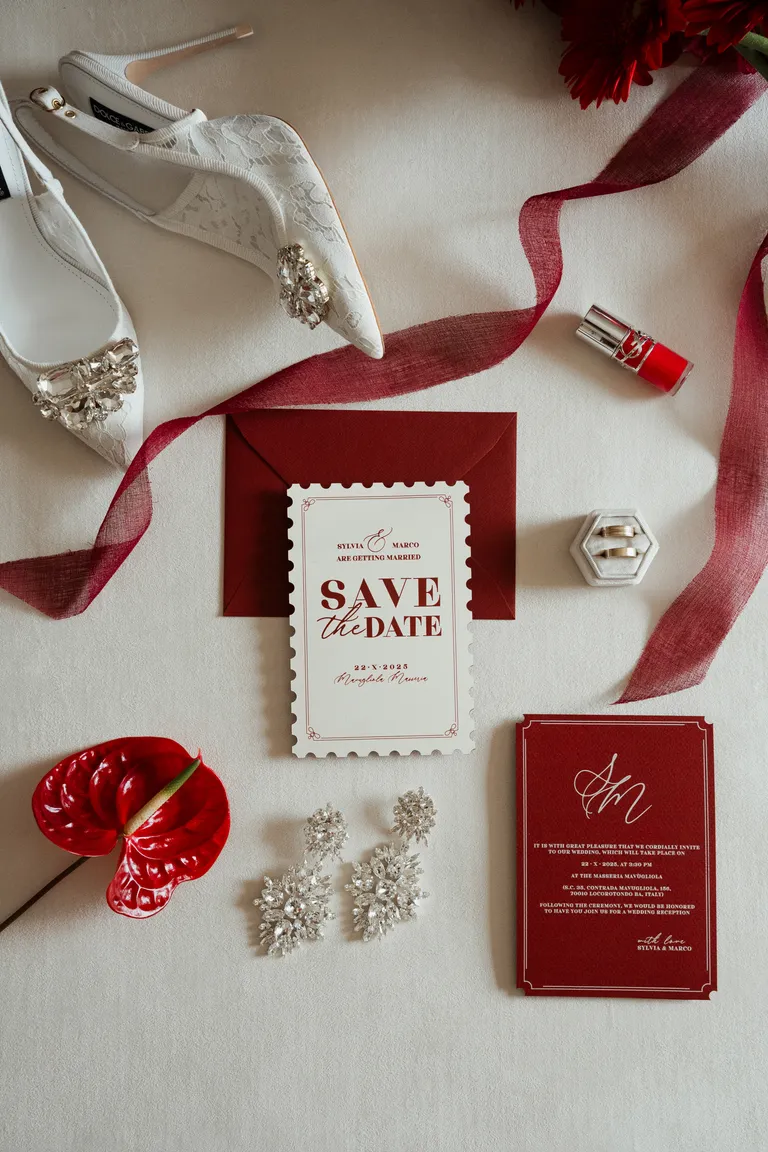 Flat-lay na jasnym blacie: zaproszenie save the date, bordowa wstążka, białe szpilki, kolczyki i czerwona szminka
