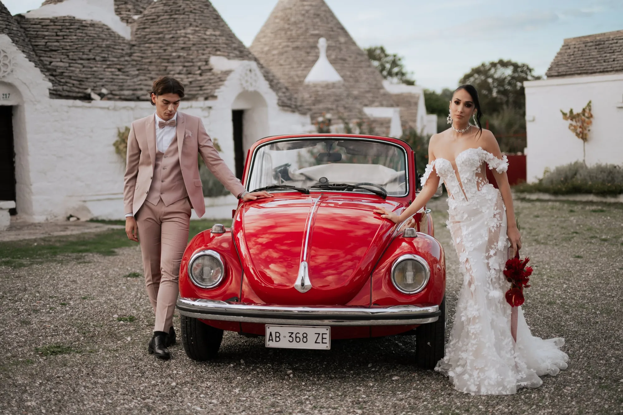 Para przy czerwonym kabriolecie retro na żwirowym placu przed białymi trulli z kamiennymi stożkowymi dachami: pan młody w pudroworóżowym garniturze, panna młoda w sukni z kwiatowymi aplikacjami i z czerwonym bukietem